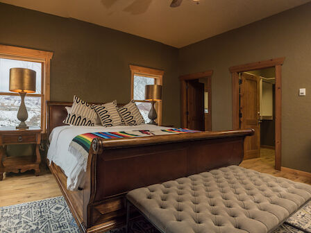 A cozy bedroom with a large wooden bed, patterned pillows, a lamp on a bedside table, a cushioned bench, and large windows with snowy views.