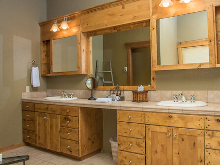 The image shows a rustic bathroom with wooden cabinetry, double sinks, large mirrors, and a well-lit vanity area. Additional items include towels and toiletries.