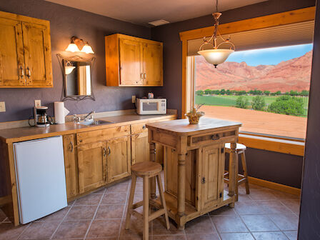A cozy kitchen with wooden cabinets, island, stools, fridge, microwave, and a stunning view of red rock formations through a large window.