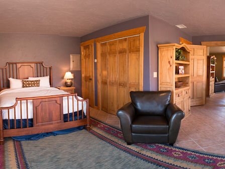 This image shows a cozy bedroom with wooden furniture, including a bed, an armchair, and a large rug. There is also a kitchenette in the background.