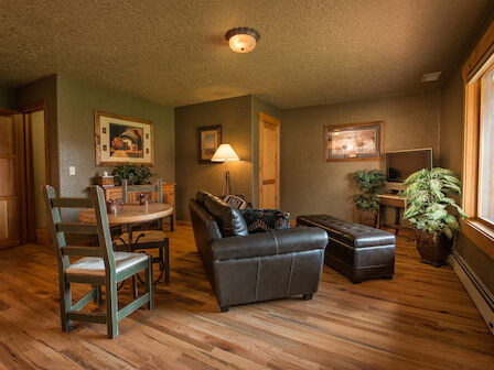 A cozy living room with a leather sofa, round dining table, chairs, artwork, plants, a TV, and large window, all on wooden flooring ending the sentence.
