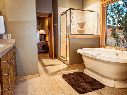 A modern bathroom features a bathtub, a brown mat, a shower, a sink, and a large window overlooking nature, with a towel hanging on the wall.