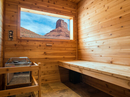 A wooden sauna room with a bench and heater, featuring a window with a scenic view of a rock formation under a bright blue sky.