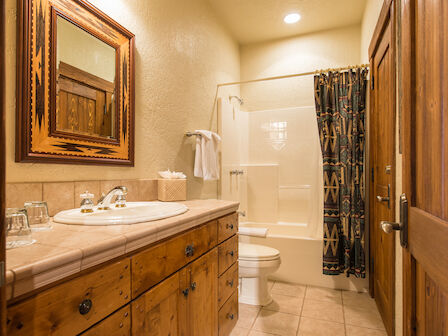 This image displays a rustic-styled bathroom with wooden cabinets, a mirror, a sink, a toilet, and a shower with a patterned curtain.