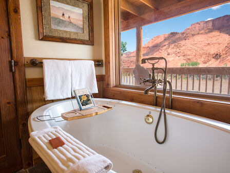 A bathroom with a tub, towels, book, art on wall, and a window view of red rock cliffs outside.