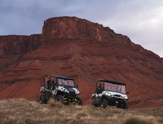 Two off-road vehicles are parked on rugged terrain with a dramatic red-rock cliff in the background.