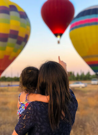 A woman holds a child, pointing at colorful hot air balloons in the sky.