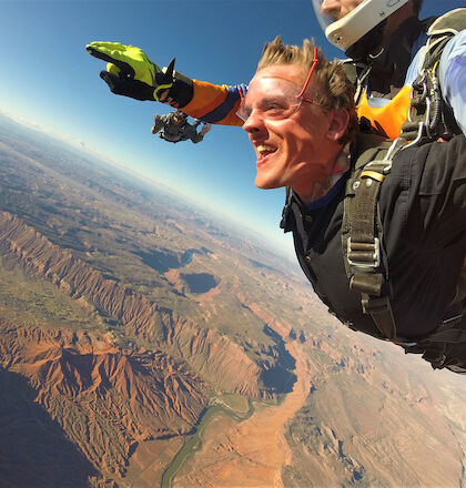 A tandem skydiving experience with two people soaring over a vast, scenic landscape featuring mountains and a winding river.