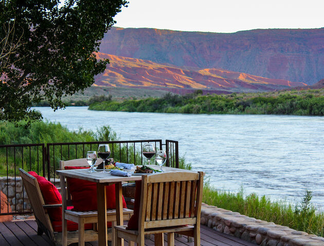 A wooden table set with wine glasses and plates overlooks a scenic river, surrounded by greenery and hills in the background.
