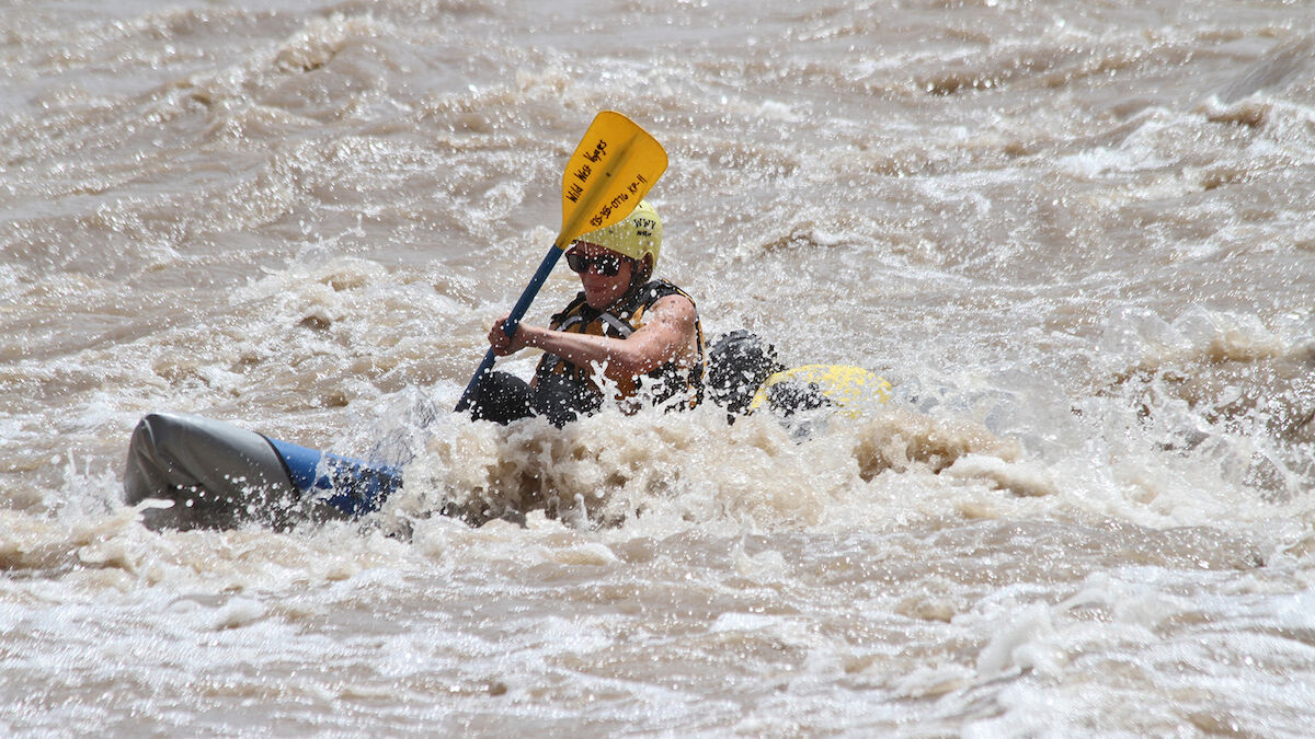 A person is kayaking through rough, frothy water, wearing a helmet and life vest, actively paddling with a yellow paddle.