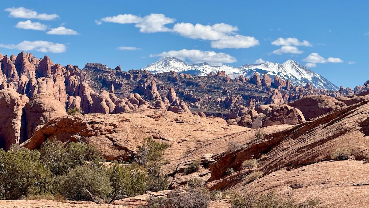 A desert landscape with red rock formations and snow-capped mountains under a blue sky with scattered clouds.