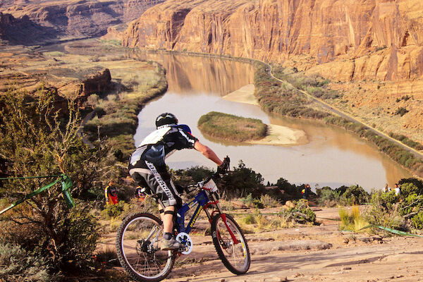 A cyclist rides down a rocky trail overlooking a river and canyon in a scenic, natural landscape.