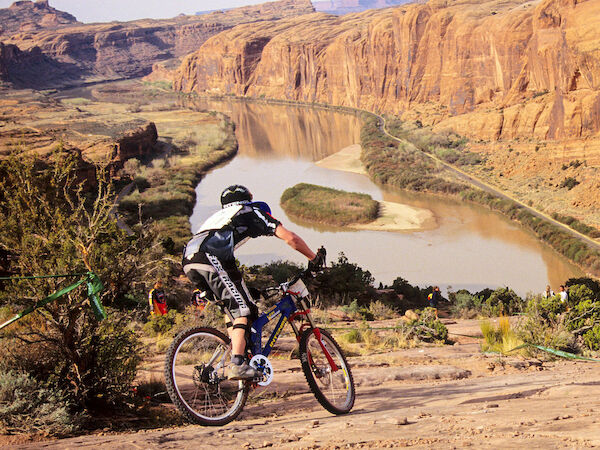A cyclist rides down a rocky trail overlooking a river and canyon in a scenic, natural landscape.