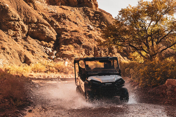 An off-road vehicle drives through a shallow rocky stream, surrounded by rugged terrain and trees under a clear sky.