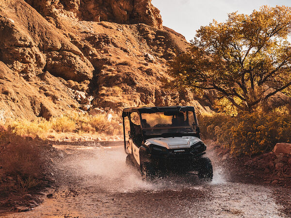 An off-road vehicle drives through a shallow rocky stream, surrounded by rugged terrain and trees under a clear sky.