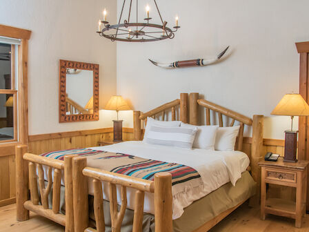 A cozy bedroom with wooden furniture features a queen-sized bed, decorative pillows, two bedside tables with lamps, a window, and rustic wall decor.