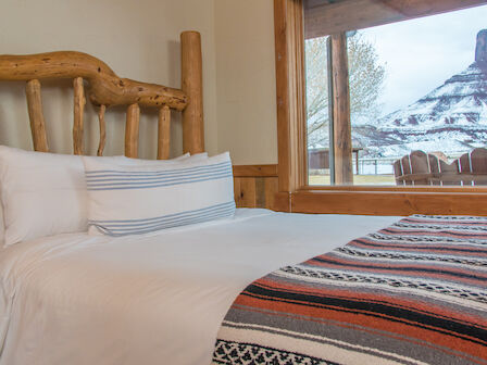 A cozy bedroom with a rustic wooden bed frame, white linens, and a striped blanket, overlooking a scenic mountain view through a large window.