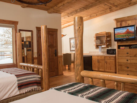 This image shows a rustic lodge-style bedroom with wooden furniture, two beds with patterned blankets, a TV, and a large window with a snowy view outside.