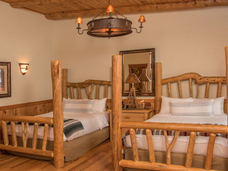 A cozy rustic bedroom features two log-frame twin beds, warm wood paneling, a chandelier, a dresser with lamps, and a framed photograph on the wall.