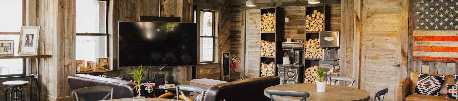 A rustic room with wooden furniture, round tables, chairs, a TV, and a wall shelf, featuring an American flag design.