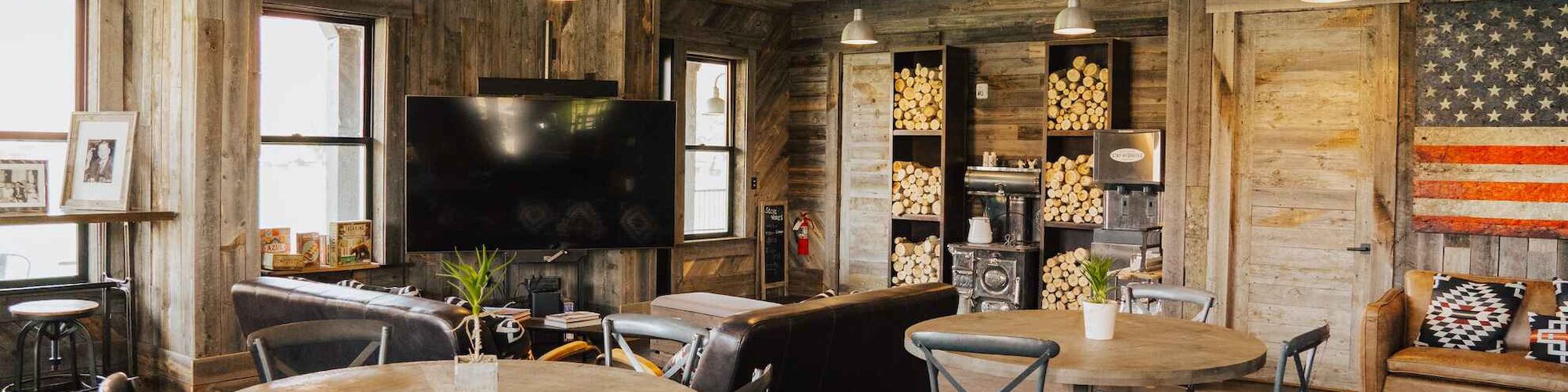 A rustic room with wooden furniture, round tables, chairs, a TV, and a wall shelf, featuring an American flag design.