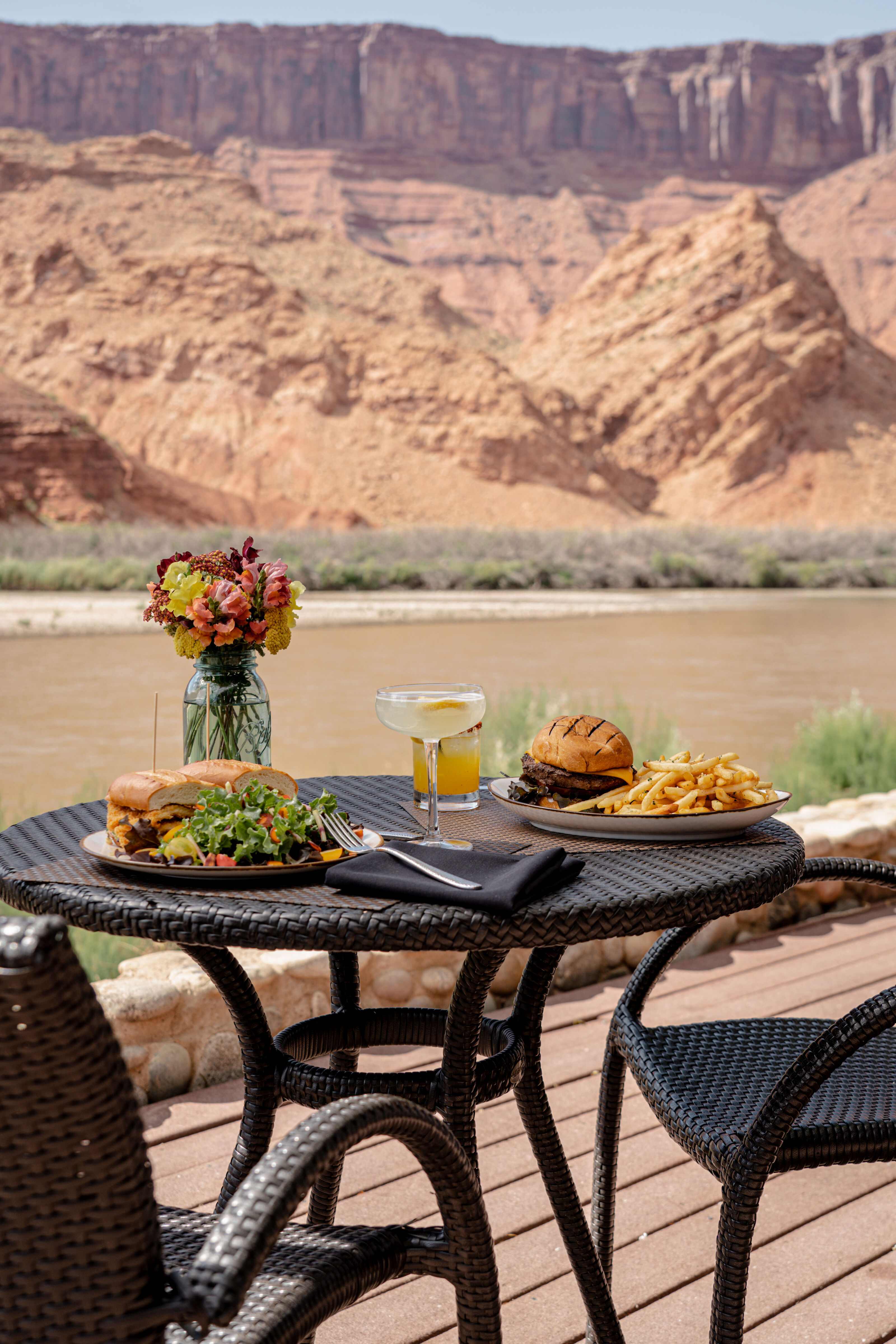 a table set for dining with food, drinks, and flowers on a patio overlooking a river and mountains
