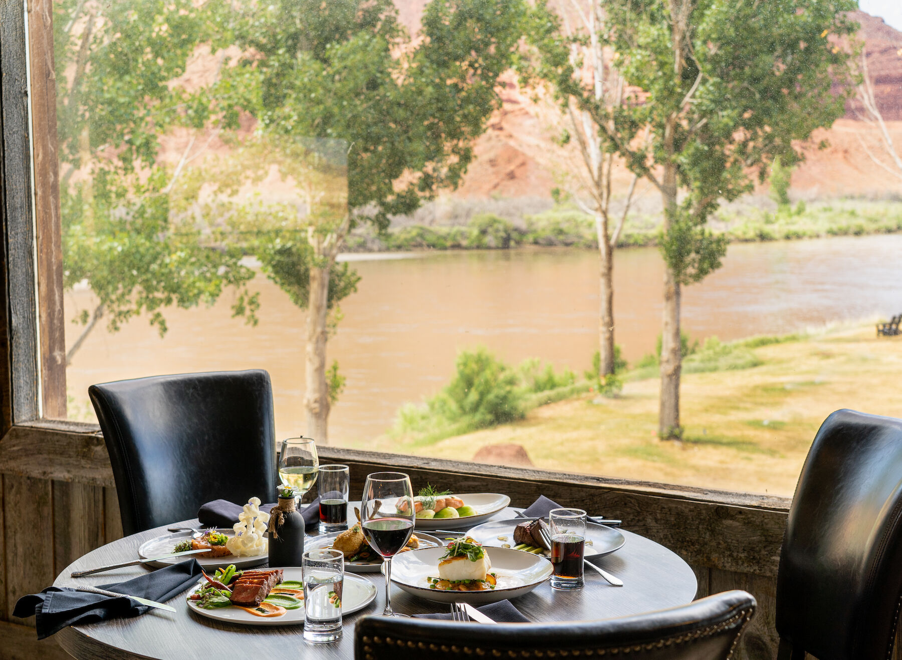 A dining table with plates of food and drinks is set by a window overlooking a river and trees in a scenic setting.