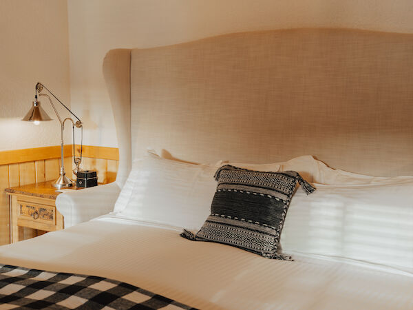 A cozy bedroom features a large bed with patterned pillows, a bedside table, and a lamp, creating a warm and inviting atmosphere.