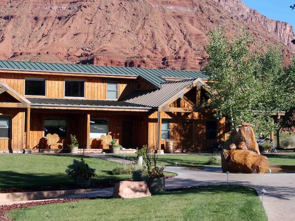 A wooden house with a green roof, surrounded by trees and a lawn, set against a backdrop of red rocky cliffs in a sunny setting.