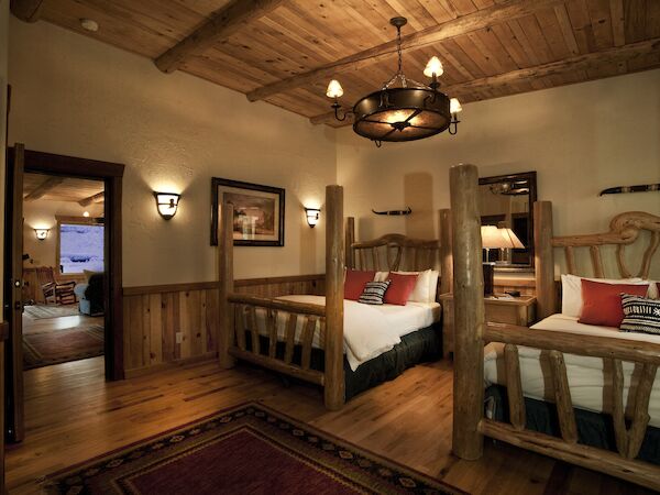A cozy rustic bedroom with two wooden beds, warm lighting, and a view into an adjoining room with a TV and fireplace.