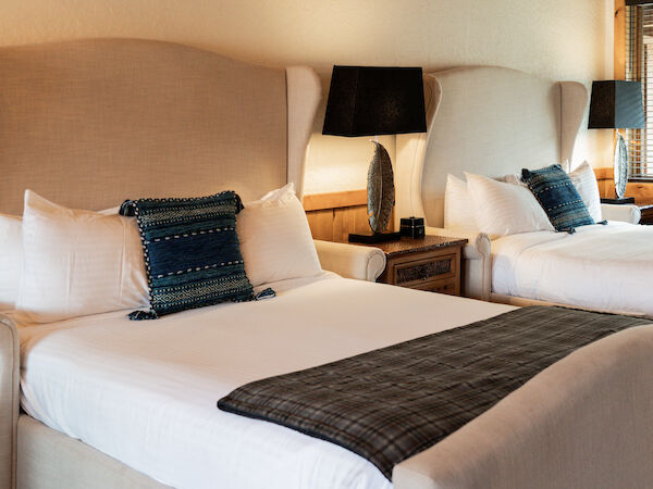 The image shows two neatly made beds with pillows and decorative cushions, accompanied by bedside lamps and a wooden table.