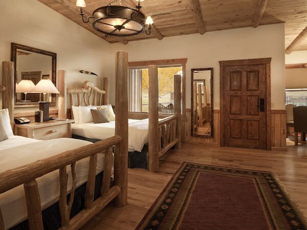 A cozy rustic bedroom features wooden furniture, two beds, a mirror, a rug, and a view into a dining area with wooden beams.
