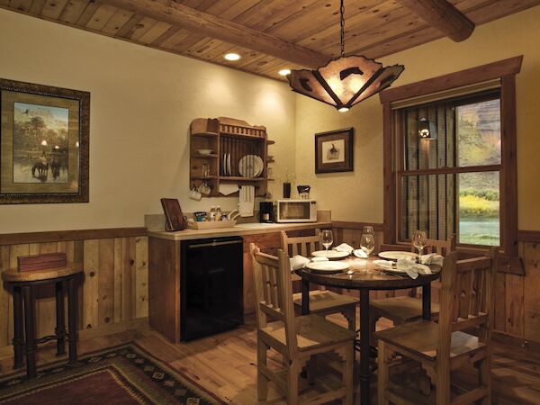 A cozy rustic dining area with wooden furniture, a table set for four, a kitchenette, and warm lighting, creating a welcoming atmosphere.