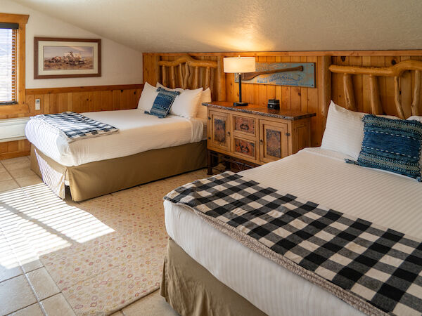 A cozy room with two beds, plaid blankets, blue pillows, wooden accents, a lamp on a rustic table, and artwork on the walls.