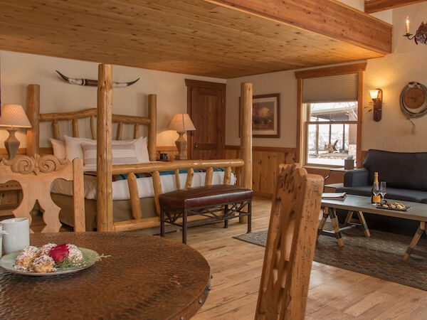 A cozy, rustic bedroom and living area with wooden decor, featuring a bed, couch, coffee table, and a small dining area with tea and snacks.