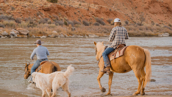Two people on horseback and a dog cross a shallow river with a rocky backdrop.