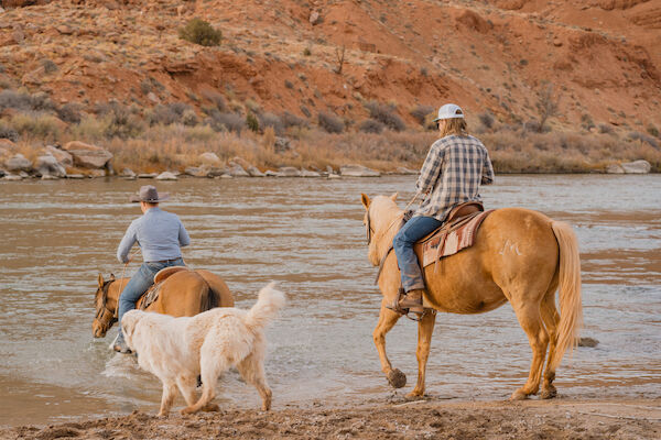 Two people on horseback and a dog cross a shallow river with a rocky backdrop.
