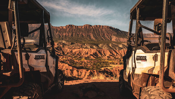 Two off-road vehicles are parked on a cliff, overlooking a desert landscape with a large mountain in the background.