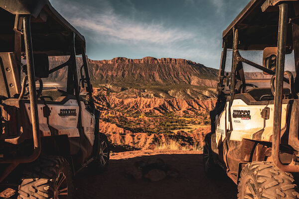 Two off-road vehicles are parked on a cliff, overlooking a desert landscape with a large mountain in the background.