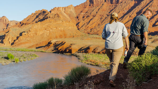 Two people walking by a river in a desert landscape, with red rock formations in the background.