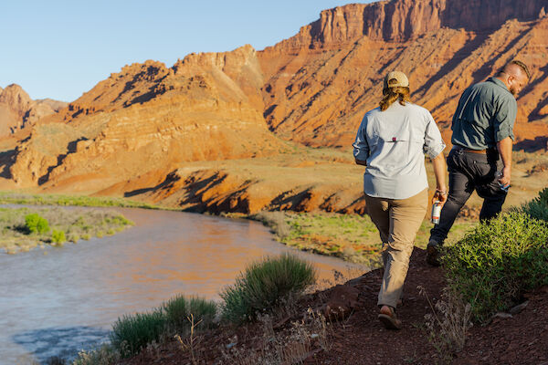 Two people walking by a river in a desert landscape, with red rock formations in the background.