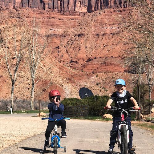 Two people are biking on a paved path with red rock cliffs and sparse trees in the background.