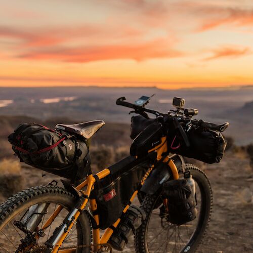 A loaded bike stands on a rocky path, set against a colorful sunset sky and expansive landscape, ready for adventure.