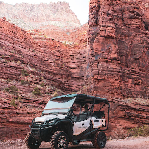 An off-road vehicle is parked on a dirt path amidst dramatic red rock formations, likely in a canyon-like landscape.