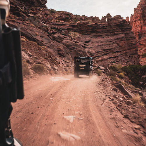 A rugged vehicle is driving on a dusty, rocky road through a canyon, surrounded by steep cliffs and sparse vegetation.