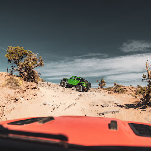 A green off-road vehicle on rugged terrain with a red vehicle's hood in view, under a clear blue sky.