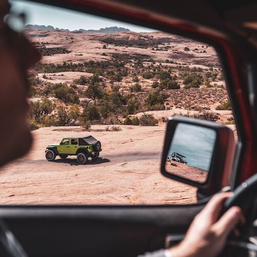 A person in a vehicle views a green off-road vehicle on a sandy landscape through the side mirror.