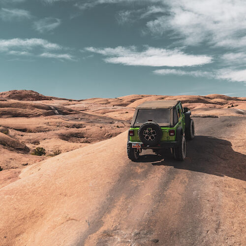 A green off-road vehicle drives on a rugged desert trail under a partly cloudy sky, surrounded by rocky terrain and distant hills.