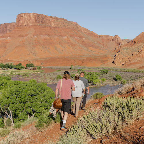 A group of people is hiking along a red rock landscape with green vegetation and a prominent rock formation in the background.