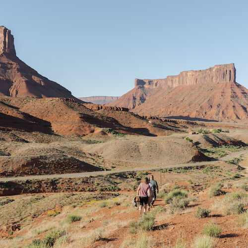 A scenic desert landscape with two large mesas, clear blue sky, and two people walking along a path surrounded by rocky terrain and vegetation.